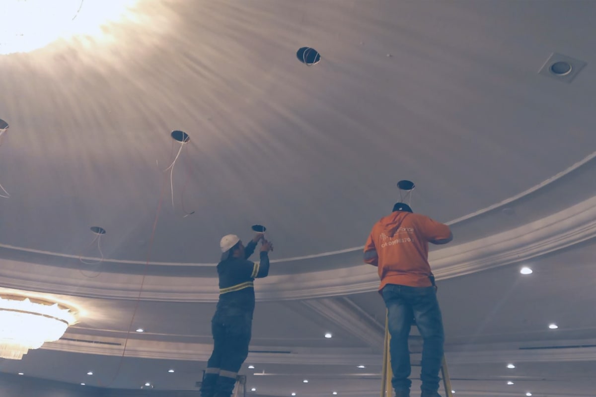 Dos hombres trabajando en las luminarias del techo de una habitación amplia.